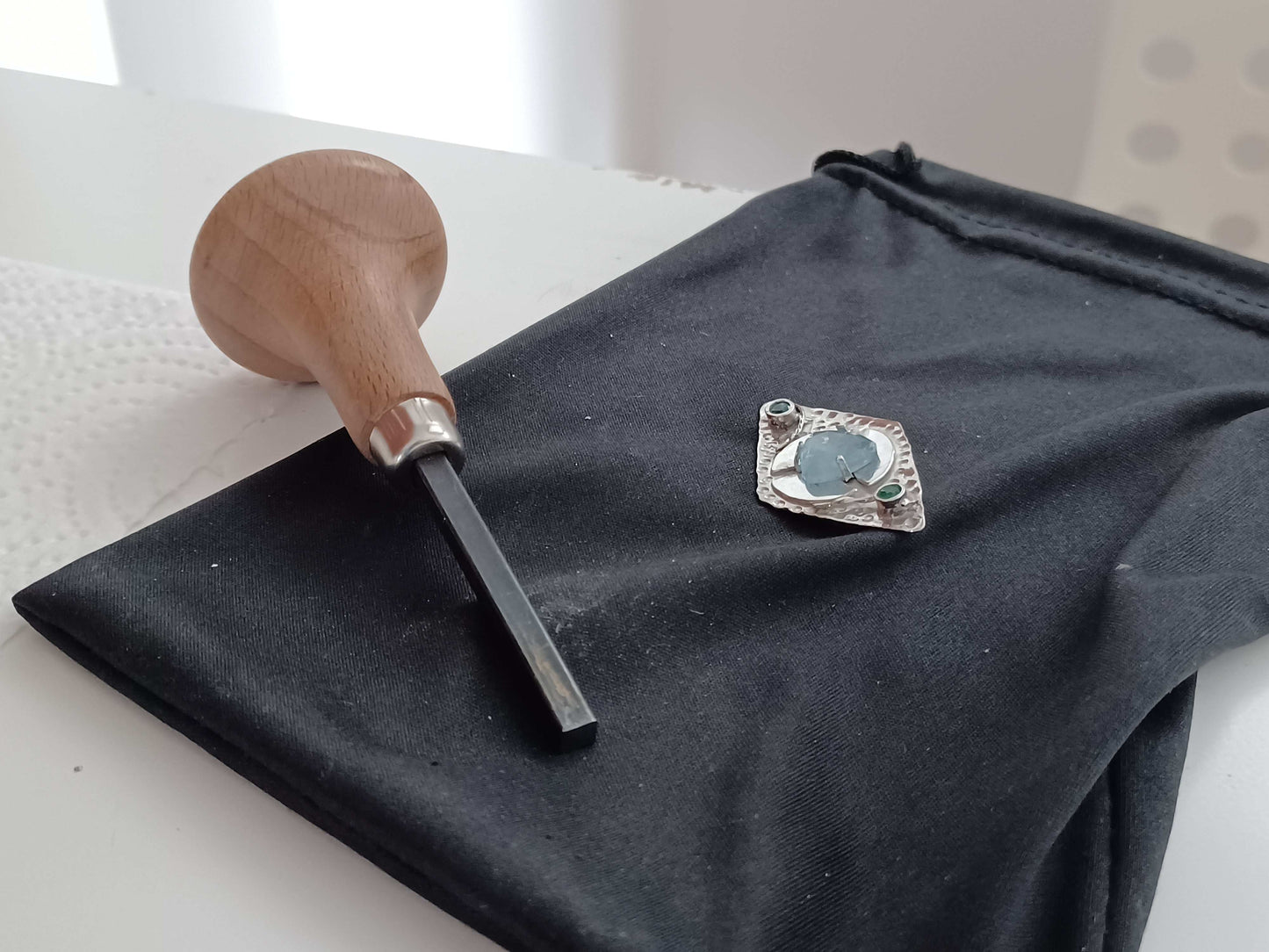 Silver pendant being made with gemstones on a black fabric surface next to a wooden stone setting tool.