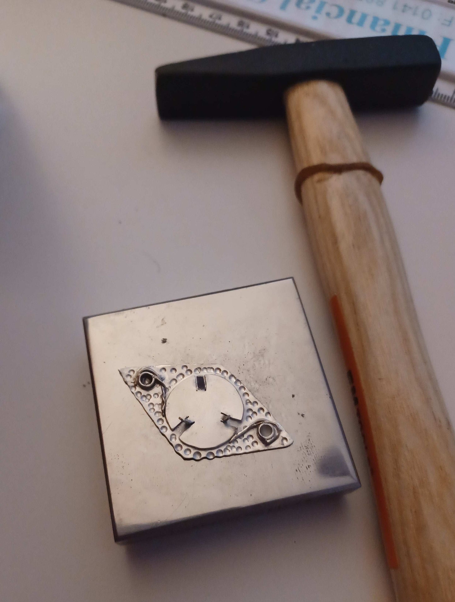 silver pendant being made on a steel block, next to a wooden handled hammer.
