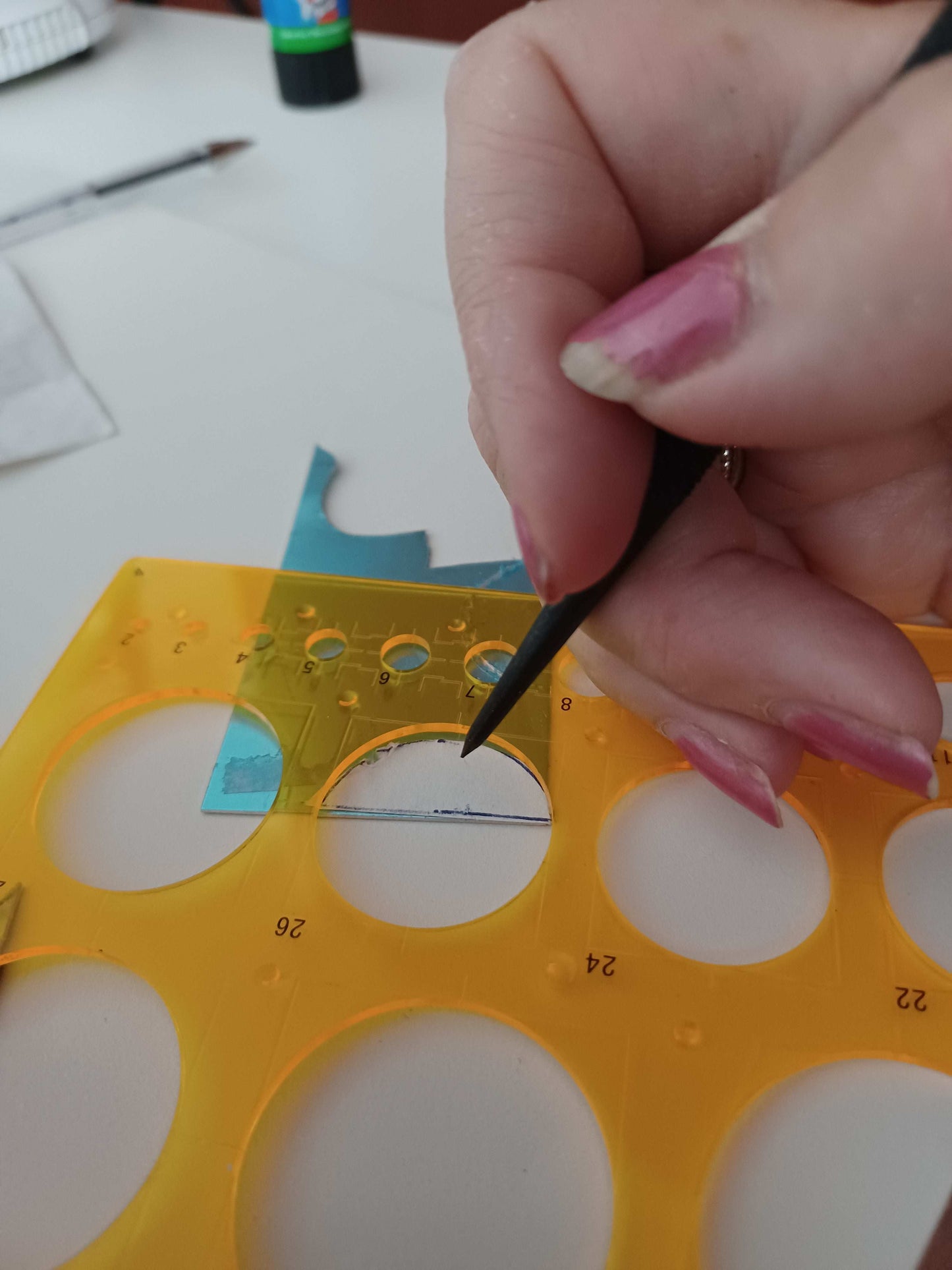 Hand holding a small scribing tool over a yellow plastic stencil with circular holes on a white background.