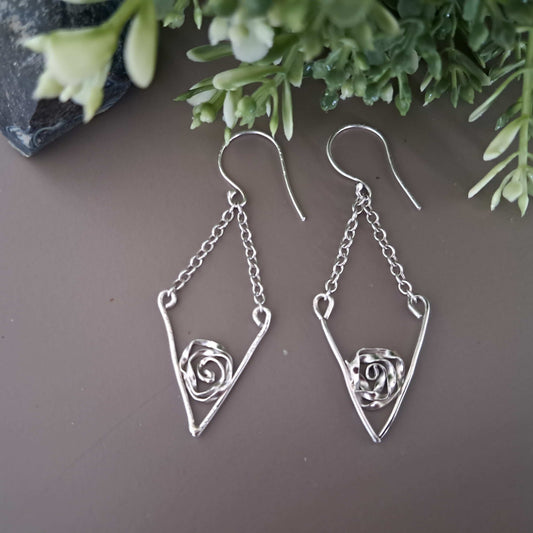 Handmade Sterling Silver v-shaped drop earrings with silver rose in the centre  on grey brown surface with green leaves in the background