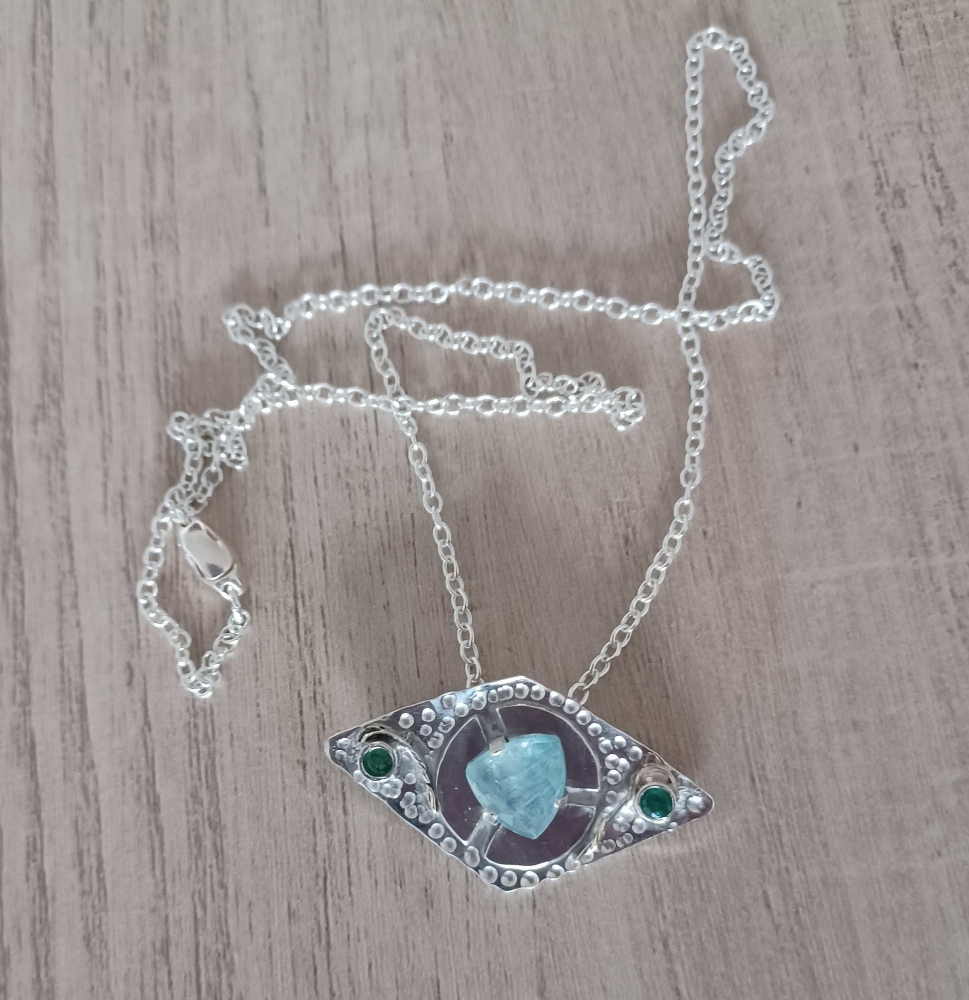 Necklace with eye-shaped pendant featuring a turquoise stone in the middle and two emeralds either side on a wooden surface