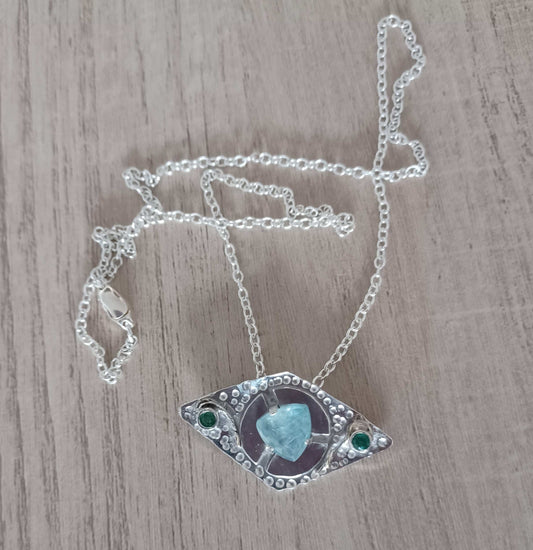 Necklace with eye-shaped pendant featuring a turquoise stone in the middle and two emeralds either side on a wooden surface